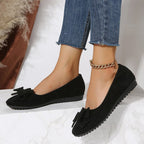 Black suede bow comfort loafers on feet with ankle chain and rubber tread sole